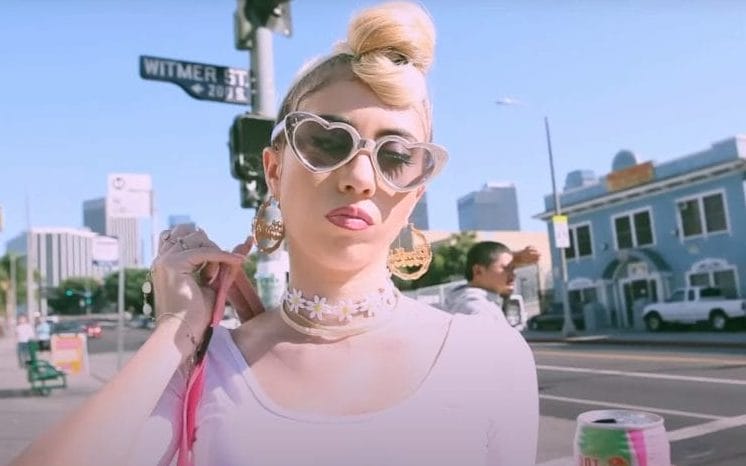 Kali Uchis wearing heart-shaped sunglasses on a sunny day in Colombia. Her hair is blonde and in a bun on top of her head. Uchis is wearing at the time trendy jewelry.